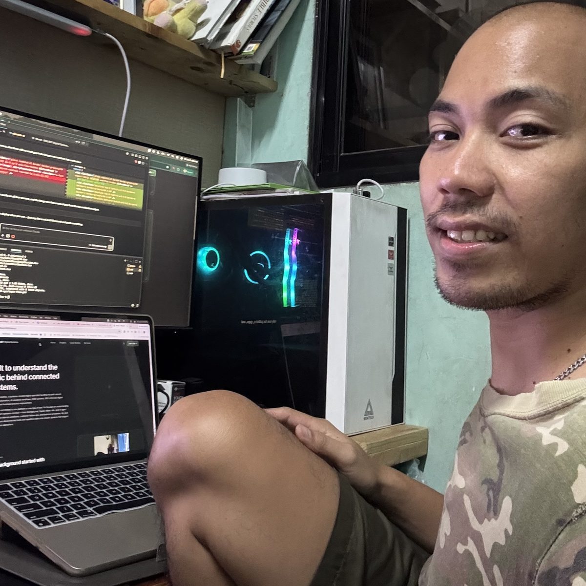 Kevin Ordoñez at his desk beside a desktop PC tower and two monitors, one showing a code editor and the other a terminal.